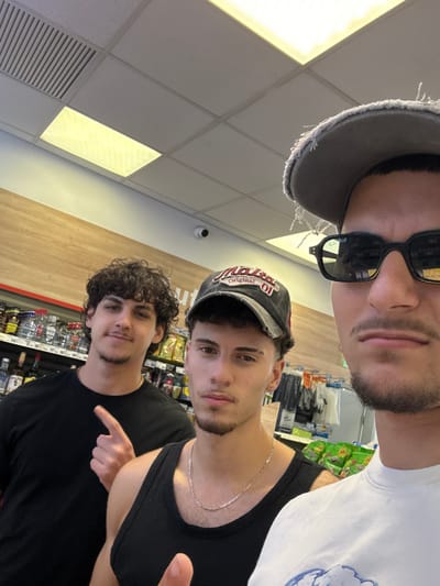 The image shows a group of three young men posing for a selfie indoors, likely in a convenience store or a similar retail environment. The lighting suggests it's daytime, possibly late afternoon or early evening given the artificial lights in the background.
The man in the foreground, on the left, is wearing a gray baseball cap with frayed edges and dark sunglasses. He has a beard and is looking towards the camera. Behind him and slightly to his right is another young man wearing a baseball cap with a logo that appears to say "Original" in red and white lettering. He has curly dark hair and is also looking at the camera. The third person, on the far right, is partially visible and appears to be wearing a white t-shirt.
In the background, shelves stocked with various products are visible, including what look like snacks in green packaging and bottles on lower shelves. The ceiling is a typical drop ceiling with fluorescent lighting panels. One of the men is pointing at something or someone to his left, and the overall mood seems casual and friendly, consistent with a group of friends taking a spontaneous photo. There are no obvious indications of the weather or time of day beyond the indoor lighting. The text "Original" is visible on the cap of the person in the middle.