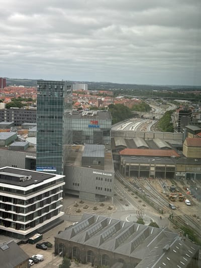 The image presents an elevated, oblique view of a complex urban environment in Aarhus, Denmark, under an overcast sky. The foreground features a modern, multi-story building with a white facade and dark-framed windows, appearing to be an office building. Adjacent to it is a large, older industrial-style building with a red-tiled roof and arched entrances, likely a former train depot or workshop, with visible train tracks in front of it.
Dominating the mid-ground is a prominent glass-fronted skyscraper, a key feature of the scene, with its reflective surface showing the sky. To the right of the skyscraper is a building with "VUE IMAX" signage. Several other lower-rise buildings with diverse architectural styles, including some with red-tiled roofs, are scattered throughout the cityscape, suggesting a mix of historical and contemporary structures.
The background reveals a complex network of railway tracks curving through the landscape, indicative of a significant transportation hub. Greenery, including trees and hills, forms a backdrop, contrasting with the urban density. Construction equipment, including yellow excavators and a white van, are visible near the railway lines, suggesting ongoing development or maintenance. A small circular traffic island with a metal sculpture is also present in the area with the railway lines. Two individuals are visible on the scene: one on a bicycle near the sculpture, and another person standing near the entrance of the older building. Several vehicles, including cars and a white van, are parked on the street and in parking areas. The overall impression is a dynamic urban area undergoing continuous change, with a strong presence of both modern architecture and essential infrastructure like railways.
