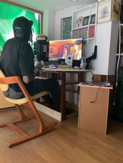 A person sits at a desk with a computer, keyboard, and monitor displaying an anime scene. A black pistol rests on a shelf above the monitor. A green screen stands behind the person. The desk appears homemade with an added shelf and supports. The person wears a black cap, black shirt, and gray pants while sitting in a wooden chair with a tan cushion. The room has a wooden floor and several framed artworks on the wall.