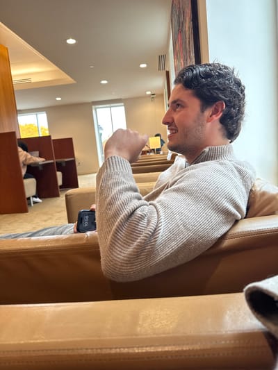 The image captures an indoor scene within a modern building in Waltham, United States, likely a university library or common area such as Bentley University. In the foreground, a young adult male with dark, curly hair, wearing a light beige ribbed knit sweater and grey pants, is seated in a light brown leather armchair. He is facing to the right, exhibiting a slight smile, and holds a black smartphone displaying "10:58" in his right hand, resting on his leg. His left hand is partially raised in a loose fist. The background features a well-lit space with multiple rows of similar light brown leather seating and individual study carrels constructed with wooden partitions. Two indistinct individuals are visible seated at these carrels further back. Large windows in the background reveal an outdoor view with visible autumn foliage. The ceiling is white, equipped with recessed lighting fixtures and a security camera. A vertical section of framed artwork, displaying abstract red and dark tones, is visible on the right wall.