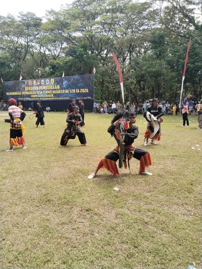 The image captures an outdoor opening ceremony on a grassy field, set against a backdrop of dense green trees, in Sragen, Indonesia. Several individuals are performing a traditional dance, likely Jaranan or Kuda Lumping, featuring elaborate horse-like props and distinctive costumes. Two central dancers are in a dynamic, crouching pose, dressed in black attire with vibrant red and yellow accents, holding their ornate props. One dancer's black top has the word "PREDATOR" visible. Other performers in similar traditional garments are scattered across the field.

In the background, a crowd of spectators observes the event, positioned beneath the trees. A large black banner stretches across the far background, announcing the "UPACARA PEMBUKAAN" (Opening Ceremony) for "TNI MANUNGGAL MEMBANGUN DESA (TMMD) REGULER KE-128 TA 2026 KODIM 0725/SRAGEN." The banner also features the slogan "TMMD SATUKAN LANGKAH, MEMBANGUN NEGERI DARI DESA" and specifies the date and location as "Sragen. 22 April 2026." Several red and white Indonesian flags on poles are visible around the banner and in the general outdoor setting. The scene unfolds during daytime, under what appears to be an overcast yet bright sky, creating a soft, even illumination.