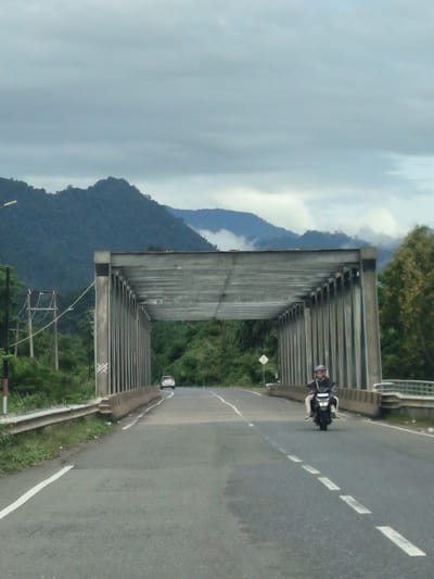 The image depicts a bridge structure over a road in Meunasahmesjid, Indonesia. A grey, overcast sky dominates the upper portion of the image, with dark, tree-covered mountains visible in the distance. A grey, concrete bridge with a metal roof extends across the road. On the road, a motorcycle carrying two individuals is approaching the camera. The rider, wearing a purple helmet, is visible, as is the person behind them. A white car is seen further down the road, also approaching the bridge. The road itself is paved and has white lane markings. On either side of the road, there are guardrails and lush green vegetation. Electrical poles with wires are present on the left side of the road, near the bridge. The overall scene suggests a rural or semi-rural setting with significant natural landscapes.