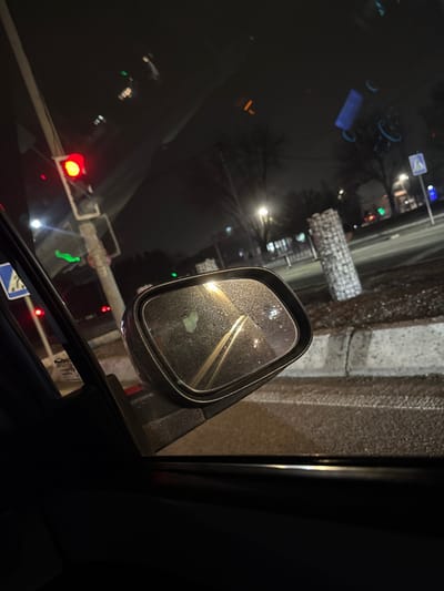 The image shows a car's side mirror with raindrops on the surface. Reflected in the mirror are blurred headlights and streetlights, creating a hazy effect. The outside environment appears dark and wet. A traffic light is visible in the lower left corner, showing a red signal. The surrounding area features buildings and road markings dimly lit. The vehicle's interior is dark.