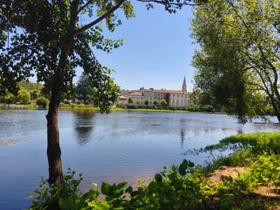 A wide shot captures a tranquil river scene in Confolens, France, with buildings and a church steeple visible on the far bank. The foreground is dominated by lush green foliage and the trunk of a tree on the left, with a sandy bank on the right. The river itself is calm, reflecting the bright blue sky and the distant architecture. Sunlight dapples through the leaves of trees framing the composition, creating a peaceful, natural ambiance. The water's surface is slightly rippled, with scattered aquatic plants adding texture. The buildings on the opposite bank appear to be a mix of residential and possibly commercial structures, with a prominent church steeple piercing the sky. The overall impression is one of serenity and natural beauty within an urban setting.