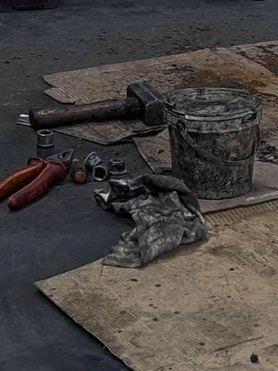 The image shows a cluttered workshop scene. A metal bucket with a handle sits next to a hammer. A soiled rag is crumpled nearby, along with several metal nuts. A pair of pliers with orange handles rests on the floor. The scene takes place on a surface covered with cardboard and a dark, possibly concrete, floor. The location is Rakovica Urban Municipality, Serbia.
