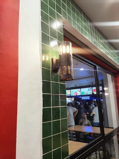 The image shows a close-up of an outdoor wall with green tiles and a white pillar. A light fixture with an open cylindrical shade is mounted on the tiled wall, emitting a warm glow. To the right, a large window reflects the interior of a building, revealing a dining area with several tables and people. The interior appears to be a restaurant or fast-food establishment, with illuminated menu boards visible. The overall setting suggests a commercial area, possibly in the city of Ariana, Tunisia, given the context. The reflection in the window is the primary indicator of human presence and activity within the establishment.