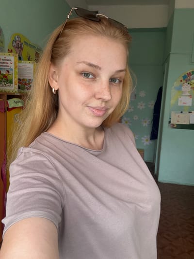 The image shows a young, fair-skinned woman with blonde hair and blue eyes. She is wearing a light purple t-shirt and a hoop earring in her left ear. Sunglasses are perched on top of her head. The background suggests an indoor environment, possibly a classroom or play area, with colorful posters and decorations on the walls. The floor appears to be tiled. There is no specific indication of the city of Vakhrushovo, Russia, within the image itself.