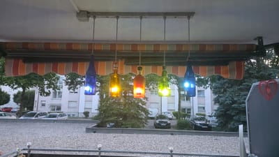 An outdoor scene shows a row of six hanging lights made from colored glass bottles. The bottles are suspended from a metal bar under an awning. From left to right, the bottles are blue, yellow, red, green, blue, and then another blue bottle to the far right. Each bottle is illuminated from within with a white light, creating colored glows that refract through the glass. The awning has horizontal stripes of brown, orange, and beige. In the background, there is a gravel-covered area, several parked cars, and a multi-story building with windows. Trees, including an evergreen tree in the center, are visible between the awning and the building. The overall scene suggests a residential balcony or patio area in Crailsheim, Germany, likely in the evening or under shaded conditions.
