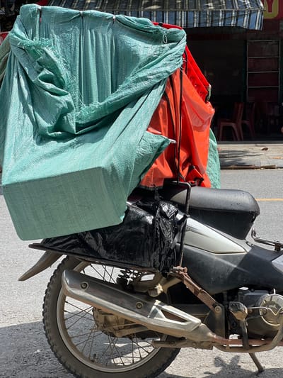A motorbike is parked on a paved street in Tân Quang Commune, Vietnam. The motorbike is loaded with a large, covered cargo. The covering consists of a teal tarp and an orange tarp. The teal tarp is draped over a bulky item, with some of its edges frayed. The orange tarp is visible underneath the teal tarp, and appears to be a separate, smaller item. A black plastic bag is tied to the back of the motorbike, below the cargo. The motorbike has a black seat, a silver and black body, and a chrome exhaust pipe. The rear wheel of the motorbike is visible, with a knobby tire. In the background, there is a building with an awning, and some red chairs. The scene is illuminated by bright sunlight, casting shadows on the street.
