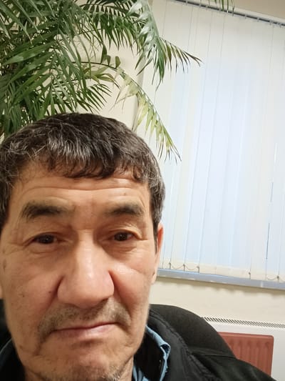 A close-up, slightly blurry selfie of an East Asian man in his late 50s or 60s is shown. He has dark, graying hair, dark eyes, and a mustache. He is wearing a dark jacket over a blue shirt. The background features a large green plant on the left, vertical white blinds on the right, and a beige wall. The scene appears to be indoors, likely in an office or home setting. There are no discernable landmarks or contextual clues indicating a specific location within Ruza, Russia. The image lacks detail to infer any specific activity or interaction.