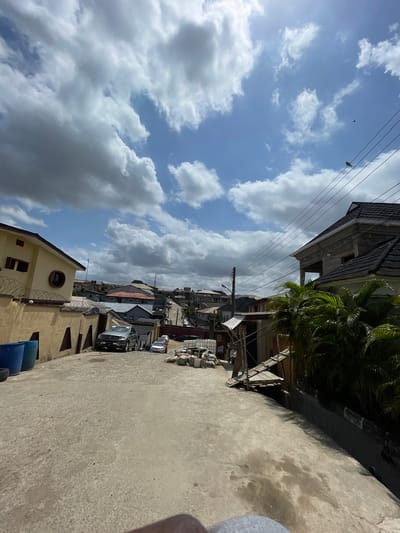 The image shows a street in Isheri, Nigeria, lined with buildings. The street is paved and appears to be moderately wide. Several parked cars are visible. The sky is partially cloudy with patches of blue sky visible. Power lines run above the street. Some buildings are under construction. There are no people visible in the image. The overall impression is a typical residential or mixed-use street scene in a developing urban environment.