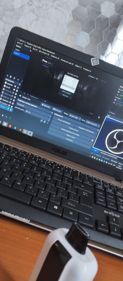 This media file captures a close-up view of a laptop screen displaying the OBS Studio software, suggesting a live streaming or recording session is in progress. The laptop itself is an ASUS model, identifiable by the logo on the bezel. The keyboard is visible in the foreground, showing a mix of Cyrillic and Latin characters, indicating a regional setting. The software interface shows various panels like "Sources," "Scene Transitions," and "Controls," with elements such as "Browser," "Device," "Image," and "Capture" suggesting different input sources are configured. The "Scene" panel highlights "Scene 2" as currently active.
In the upper right corner of the screen, a small QR code is affixed to the laptop bezel. The software also displays options for "Start Streaming," "Start Recording," and "Virtual Camera." Further to the right, a YouTube Live Control Panel is partially visible, showing elapsed time and other streaming metrics. The overall scene suggests someone is preparing for or actively engaged in broadcasting content online, potentially gaming or commentary given the software's typical use.
The lighting appears to be artificial, consistent with an indoor setting, and the time of day is not discernible. The background behind the laptop features a hexagonal tiled wall, possibly in a modern interior. The presence of a white and black electronic device in the extreme foreground, partially obscuring the keyboard, adds another object to the scene, but its function is unclear. No people are directly visible in the frame. The location context of Bamboura, Abkhazia, is provided, but there are no explicit visual cues in the image to confirm this specific location.