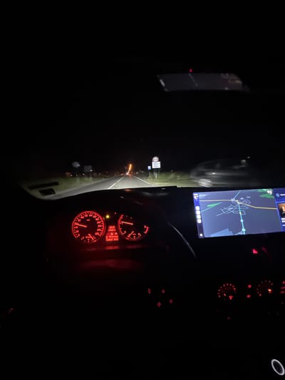 The image shows the interior of a car at night. The dashboard gauges are illuminated in red. A navigation app is displaying a map. The location is Mogila, Bulgaria as it is visible on the navigation system. The car is driving on a road that goes through a dark environment with visible street lights on the side. The driver is navigating based on the app.