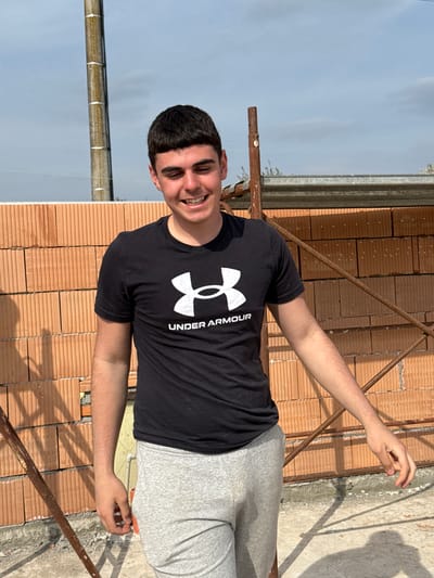 A young man wearing a black Under Armour t-shirt and gray sweatpants stands on a construction site in Goliam Dol, Bulgaria. He is smiling and has his arms outstretched. The background consists of brickwork, scaffolding, and a partially cloudy sky.