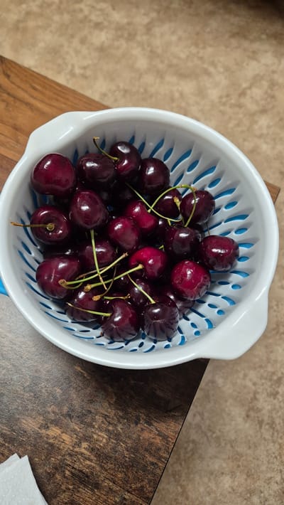 A white colander filled with dark red cherries sits on a wooden table. The cherries are ripe and glossy, with some stems still attached. The colander has a blue patterned insert. The wooden table has a visible grain and is slightly worn. The background appears to be a neutral-colored countertop and wall. There are no people or animals visible in the image, and no actions or interactions are taking place. The scene is static and appears to be an indoor setting. The location in the city of Miltenberg, Germany, cannot be determined from the visual information provided in the image.