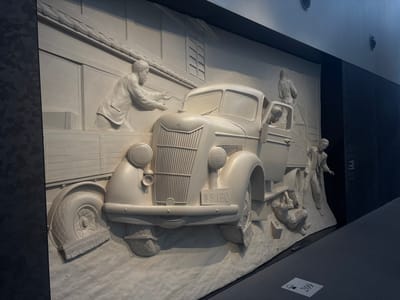 This image depicts a large, three-dimensional bas-relief sculpture, likely in a museum or exhibition space. The sculpture is rendered in a light-colored material, possibly plaster or stone, and is mounted on a dark wall. The scene portrays a historical moment involving a vintage truck and several figures.

The central focus is a light-colored truck, reminiscent of a mid-20th-century model, with a prominent grille and rounded headlights. A license plate on the front reads "S.3.180" in English and possibly Japanese characters. The truck has an open cab with figures inside and around it.

On the left side of the sculpture, a man in a suit is leaning over the truck's bed, appearing to be loading or unloading something. His hands are extended towards the truck.

In the truck's cab, a figure is visible, looking outwards. Another figure stands behind the truck, facing away from the viewer, possibly directing or overseeing the operation.

To the right of the truck, another man in a suit is leaning down, appearing to be working on the truck's tire or wheel, with a small suitcase next to him.

The background of the sculpture depicts architectural elements, possibly a building with a tiled roof and windows, suggesting an urban or industrial setting. The ground beneath the truck and figures is rendered in a textured, uneven manner, suggesting a natural or rough terrain.

The sculpture is illuminated by artificial lighting, casting shadows that enhance the three-dimensional effect and highlight the details of the figures and the vehicle. The overall presentation is clean and well-maintained, indicative of a public exhibition.

The provided hint that this is in Nagoya, Japan, suggests this sculpture might be related to the industrial history of the city, possibly commemorating transportation or early automotive development in Japan. The license plate format, with English characters and numbers, might be a stylistic choice to represent a historical period or a specific event.