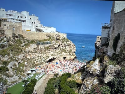 The image depicts a coastal scene with a beach crowded with people sunbathing and swimming. Buildings, some white and modern, others made of stone, are situated on cliffs overlooking the sea. A small white boat is visible in the turquoise water. Lush green vegetation grows on the rocky slopes. The sky is a clear, vibrant blue. The scene is a busy beach resort. This image is not of Porrentruy, Switzerland.