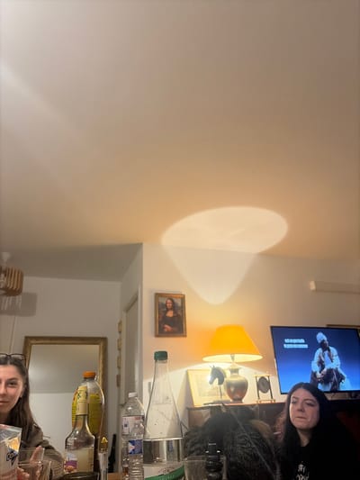 A living room scene features two women. One woman is lying on a couch watching television while the other is seated at a table. The table is cluttered with bottles. The room also includes a table lamp, a framed portrait of Mona Lisa, a mirror, and a speaker mounted on the wall. The overall composition has a casual and lived-in feel.
