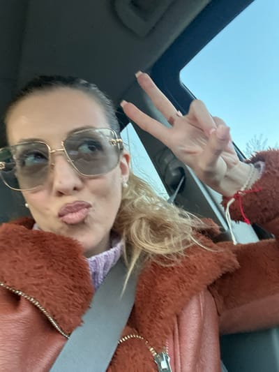 A woman is taking a selfie in what appears to be a car. She is wearing sunglasses and a brown fuzzy jacket, and she is making a peace sign with her hand. The car has a sunroof. The image does not contain any information to indicate the location as the city of Basildon, United Kingdom.