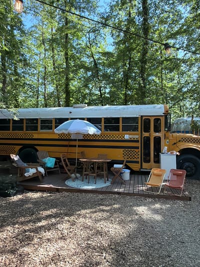 The image shows an outdoor seating area set up on a wooden deck next to a converted yellow school bus. The bus is parked in a wooded area with tall trees and dappled sunlight. Several wooden chairs and a table with an umbrella are arranged on the deck. Two folding chairs are positioned in front of the bus. A small white cooler or refrigerator is visible next to the bus. In the background, another similar bus or vehicle can be partially seen. The ground around the deck is covered with wood chips. The scene appears to be a recreational or glamping setup. The city of Cavriglia, Italy, is not depicted or suggested by any elements in the image.