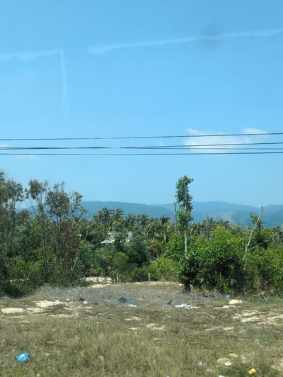The image shows a view from inside a vehicle, looking out towards a landscape in Tuy Phong 4, Vietnam. On the left side of the image, there is a window with a clear blue reflection. Power lines run vertically in the middle ground. Beyond the power lines, there is a landscape with lush green vegetation, including trees and possibly coconut palms, against a backdrop of distant blue mountains. The foreground on the right side is a patch of dry, grassy land. There is some trash visible on the ground.