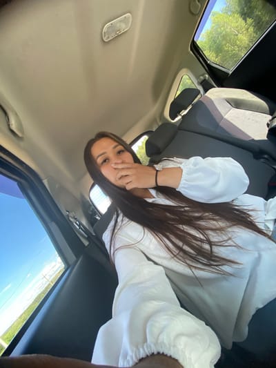 A young woman with long, dark hair is sitting in the back seat of a car, looking at the camera. She is wearing a white blouse with puffed sleeves and a bracelet on her left wrist. Her right hand is covering her nose. The interior of the car is visible, including the headrests of the seats and the ceiling with a light fixture. Outside the windows, trees and a bright blue sky can be seen. The image appears to have been taken during the daytime. The scene is likely within or near the city of Isyangulovo, Russia, based on the provided context, and depicts a person inside a vehicle during daylight.