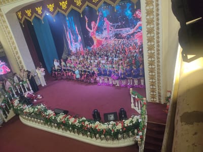 The image depicts a formal indoor event, likely a children's dance performance or awards ceremony, taking place on a large stage in Qaskeleñ, Kazakhstan. The stage is adorned with a large screen displaying colorful imagery of dancers, and is framed by ornate, gold-embellished curtains. A large gathering of children, dressed in various colorful costumes, are assembled on the stage. Many of the children are holding trophies, suggesting an awards presentation. The foreground features a richly decorated red carpeted floor and a white railing adorned with floral arrangements. In the background, several adults, presumably performers or organizers, are also visible. The overall atmosphere appears festive and celebratory.