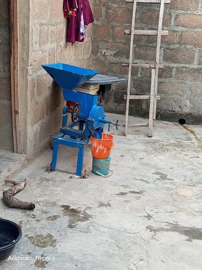 The image shows a blue electric grinder machine with a metal frame and legs. It is sitting on a concrete surface, adjacent to a brick wall. An orange bucket and a blue container are located next to the grinder. A wooden ladder is leaning against the wall. A piece of wood lies on the ground, and a partially visible black bucket is in the bottom left corner. The scene is in a residential area, likely in a backyard or a small courtyard. The location is in Ilobu, Nigeria.