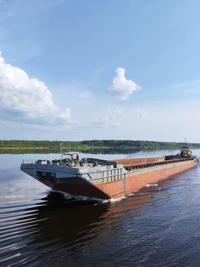 A barge navigates a river under a blue sky with scattered clouds. The barge is predominantly gray and red, creating ripples in the dark water. A line of green trees is visible along the shoreline in the background. The image does not contain any information about people or objects being in the city of Kochino, Russia.