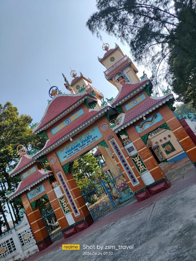 The image shows the ornate entrance gate of a Buddhist temple in Gò Dầu, Vietnam. The gate is elaborately decorated with traditional Vietnamese architectural elements, including multi-tiered, tiled roofs, colorful murals, and intricate carvings. The dominant colors are red, yellow, and blue, with accents of green and white. The gate features large, arched openings with blue metal bars, suggesting it leads into a temple courtyard. Above the archways, Vietnamese inscriptions are visible, indicating the temple's name and possibly its affiliation. Flanking the central entrance are two taller structures resembling pagodas or towers, topped with decorative spires. The entire structure is set against a clear blue sky, with lush green trees visible on either side, suggesting a natural or semi-rural setting. The ground in front of the gate is paved with concrete. The perspective of the photo is angled upwards, emphasizing the grandeur of the temple entrance. A watermarked date and time "2026.06.24 07:30" and "realme Shot by zsm_travel" are present in the lower right corner. There are no people visible in the image.