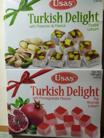 The image displays two boxes of Turkish Delight. The top box, featuring a green ribbon graphic, is white with green text that reads "Turkish Delight with Pistachio & Peanut". It also shows images of pistachios and peanuts, and the product itself as small, cubed confectioneries dusted with powdered sugar and containing green pistachios. The bottom box, decorated with a red ribbon graphic, is also white with red text that reads "Turkish Delight with Pomegranate Flavour". This box displays images of a whole pomegranate and a halved pomegranate, along with red-colored cubed Turkish Delight. The brand name "Usaş" is visible on both boxes, accompanied by weight information in ounces and grams. The setting is a plain surface, potentially a countertop or display area, with a neutral background. No specific location in Bilajari, Azerbaijan, is depicted or implied.
