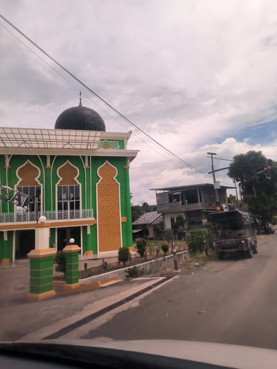 This image captures a daytime street scene in Meunasah Kumbang, Indonesia, offering a view from inside a vehicle. The primary subject is a vibrantly colored mosque on the left side of the frame. The mosque features a bright green facade adorned with intricate golden-yellow Islamic geometric patterns around its arched windows and as textured panels. A prominent dark, tiled dome, topped with a finial, crowns the building. Below the dome, a white balcony with a patterned railing extends across the front, and a modern translucent roof covers a portion of the entrance. The mosque's entrance also features decorative green and yellow pillars.
On the right side of the paved road, a dark-colored Mitsubishi truck is parked, facing away from the viewer. The word "MITSUBISHI" is clearly visible on its tailgate, with "UNISIN" written below it. Behind the truck, a two-story building in a state of partial construction or disrepair, made of wood and concrete, stands out. Further back, another smaller house is partially visible. Utility poles and a network of overhead wires crisscross the cloudy sky, which is a mix of white and grey, with some patches of blue showing through. A pole near the truck holds amber lights, possibly traffic signals or warning lights.
Green foliage from trees and smaller shrubs is scattered throughout the scene, adding natural elements to the urban landscape. The bright illumination suggests it is daytime. The image provides a candid glimpse into a local street environment, with no discernible human activity beyond the implied presence of the photographer in a vehicle.