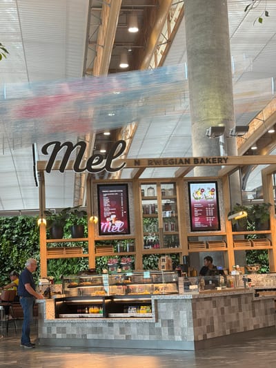 The image depicts the interior of a bakery and cafe, likely "Mel Norwegian Bakery," located in Gardermoen, Norway. A man in a dark blue shirt and jeans stands at the counter, appearing to interact with a point-of-sale system. To his right, a staff member in a black shirt is visible behind the counter. The cafe features a display of baked goods under glass, a wall of green plants, and wooden shelving units. Above the counter, a large illuminated sign reads "Mel," with "N RWEGIAN BAKERY" also visible. Two digital menu boards are mounted on the shelving. The ceiling is a complex structure of beams and panels, with decorative strands of fabric hanging from above. The overall environment suggests a modern and well-lit commercial space.