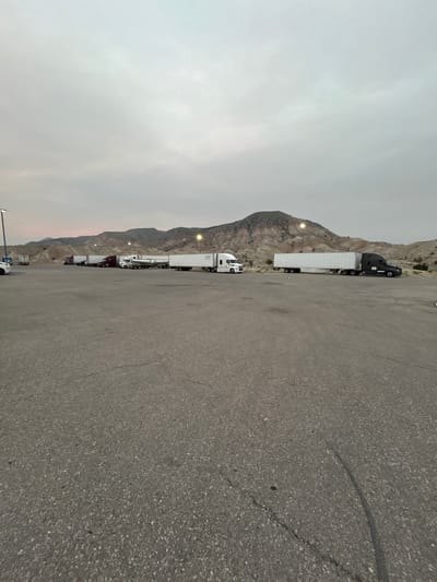 The image captures a truck stop or rest area in a mountainous, arid region, possibly in the American West, given the location context of Salina, USA. The scene is dominated by a row of semi-trailer trucks parked on an asphalt lot. The trucks are of various colors, with the most prominent being a black tractor pulling a white trailer, followed by several other white trailers and tractors in shades of white and dark red. The sky is overcast with a hazy, light grey appearance, suggesting it might be early morning, late afternoon, or a day with smoke from wildfires. The mountains in the background are rugged and appear to be made of rock, with visible layers and striations. There are streetlights along the edge of the parking lot, some of which are illuminated. The asphalt shows signs of wear, with visible cracks and tire marks. The overall mood is functional and utilitarian, typical of a rest stop for long-haul truckers. There is no visible text in the image.