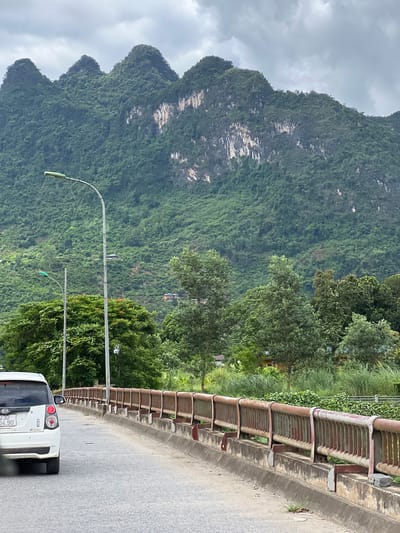 The image shows a bridge with a white Kia car driving on it, with a mountainous landscape in the background. The bridge has a rusty metal railing and concrete barriers. The mountains are covered in lush green vegetation, with some rocky outcrops visible. The sky is overcast. In the foreground, the road is asphalt, and there is some grass growing along the side of the bridge. The car has a license plate that reads "23D 04.90". There are streetlights on the bridge. The general location is within Hà Giang 2 Ward, Vietnam, given the style of the mountains and the vehicle.