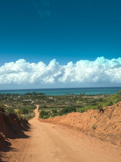 The image depicts a dirt road winding through a landscape under a bright blue sky. The road is made of reddish-brown earth and is flanked by sandy embankments on either side, with sparse green vegetation growing on them. In the distance, the road leads towards a coastal area where the turquoise ocean meets a hazy horizon. Along the coastline, there are clusters of trees and some buildings, suggesting a small settlement or village. Large, white cumulus clouds fill the upper portion of the sky, creating a stark contrast with the deep blue. The overall impression is one of a remote, rural, and possibly tropical environment.
