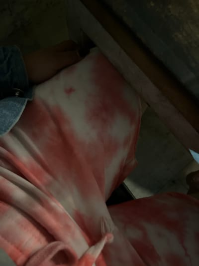 The image shows a close-up view of someone wearing a pink and white tie-dye hoodie, partially obscured by what appears to be a dark, possibly wooden or metallic, structure. A small portion of a blue denim jacket is visible at the lower left. The lighting is dim. No information suggests any connection to Desa Delod Peken, Indonesia. The image only focuses on clothing and immediate surroundings.