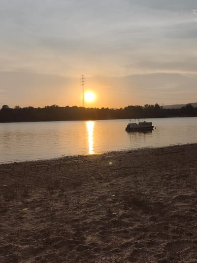 This media file depicts a sunset over a calm body of water, likely a river or lake, in Dunakeszi, Hungary. The foreground is a sandy shoreline with scattered pebbles and sparse vegetation. The water reflects the warm, golden hues of the setting sun, creating a bright, shimmering path across its surface.
In the middle ground, a pontoon boat is moored on the water, appearing somewhat weathered. The far bank of the water is lined with a dense band of trees, silhouetted against the sky. A tall, lattice-like electricity pylon stands prominently on the horizon, with power lines extending from it. The sun itself is a bright orb, partially obscured by the pylon and casting a warm glow across the sky. The sky is a gradient of pale orange and gray, with some faint clouds. The overall mood is peaceful and serene, characteristic of a quiet evening by the water. The time of day is clearly sunset. There is no visible text in the image.