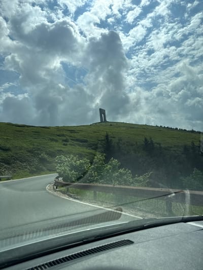 The image depicts a mountainous landscape near Hristo Danovo, Bulgaria, captured from inside a vehicle. A prominent, dark, arch-like or U-shaped structure stands atop a large green hill in the mid-ground, silhouetted against a partially cloudy sky. The sky features a mix of bright blue areas and dense white to grey cumulus clouds, with multiple airplane contrails visible. An asphalt road curves left in the foreground, its edge marked by a white line, and a metal guardrail runs along the right side of the road. The hillsides are covered in green grass and scattered trees and bushes. The lower portion of the image shows the vehicle's dashboard and a reflection on the windshield. No people or animals are present.