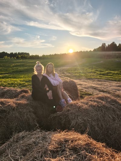 The image portrays two women seated on large, round hay bales in a rural setting during sunset, likely in Стеклянный, Russia. The low-lying sun, a bright golden orb, is visible on the right side of the horizon, casting a warm, golden light and producing noticeable lens flares across the scene.

In the foreground, a prominent pile of dry, golden-brown hay bales occupies the lower portion of the frame. The woman on the left, with light-colored hair tied in a bun, is dressed in a dark, long-sleeved jumpsuit. She looks directly at the camera with a neutral expression, her hair beautifully backlit by the sun. Beside her, the woman on the right, with long, light brown hair, wears a light pink hooded sweatshirt and light-colored shorts. She is turned slightly to her left, away from the camera, and offers a gentle smile. Her legs are crossed, and she wears athletic shoes with pink details.

Beyond the women and hay bales, a green field extends into the mid-ground, featuring a faint dirt path that curves through it. A dense line of dark green trees forms the distant horizon, indicating the edge of a forest. The sky above is a blend of soft blue, light orange, and yellow hues near the sunset, transitioning to a clearer light blue higher up, dotted with delicate, wispy white clouds. The overall atmosphere is serene and warm, typical of a late afternoon or early evening in the countryside. No visible text is present in the image.