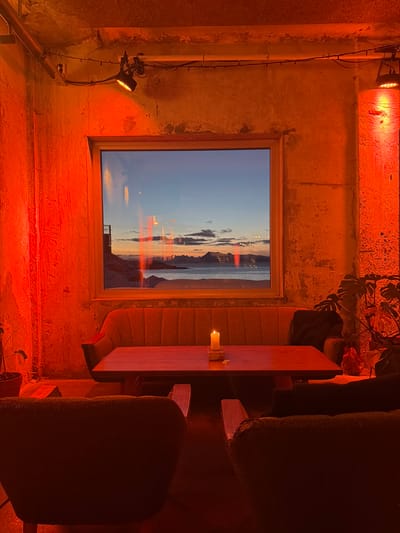 A dimly lit, orangey-red room features a window with a beach sunset view. The walls are concrete and somewhat distressed. A small lit candle sits on a narrow wooden table, next to a padded bench seat. Two chairs, upholstered in a dark fabric, are partially visible. A spotlight attached to a metal pipe illuminates the wall to the left of the window. A plant sits in the top right of the frame.