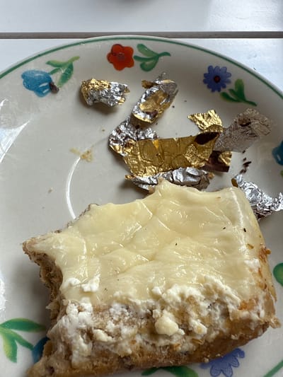 A slice of bread with a creamy white topping is on a plate with a floral design. There are pieces of gold and silver foil scattered on the plate. The plate is white with green trim and painted blue and orange flowers.