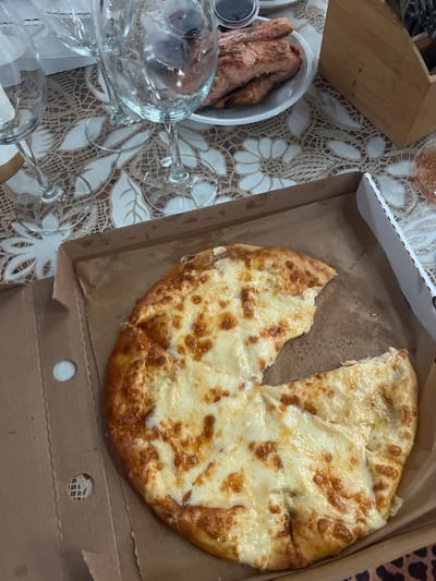The image displays a tabletop scene featuring various food items and drinkware, indicative of a casual meal or gathering, potentially in an indoor setting.

The central focus is a large, round, cheesy flatbread, resembling a Khachapuri, presented in a brown cardboard take-out box. A single slice has been removed from the flatbread, suggesting it has already been partially consumed. The cheese topping is melted and has areas of golden-brown caramelization, and the crust is also golden. Grease marks are noticeable on the bottom of the cardboard box, beneath the flatbread.

To the left of the flatbread, a white bowl contains several pieces of what appear to be fried chicken or similar cooked meat, characterized by a crispy, dark brown skin. Next to this bowl is a small, clear cup holding a dark dipping sauce. Scattered across the foreground and left side are multiple clear glass wine glasses or tumblers, some appearing empty, others possibly containing ice or residual liquid.

The table is covered by a decorative tablecloth featuring a beige background with an intricate white pattern of floral or leafy motifs. A wooden cutting board or tray is partially visible in the upper-left quadrant, while a small section of a leopard-print object can be seen in the upper-right corner.

No people are visible in the frame, and there is no discernible text on any of the objects. The informal setting and food choices could be consistent with the provided location context of Zima Yuzhnaya, Russia, suggesting a casual dining experience.
