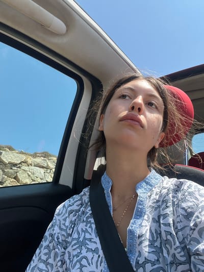 The image depicts a female individual seated within the passenger compartment of a vehicle, consistent with a convertible or vehicle with a large sunroof, under a clear blue sky. The scene is located in Agios Ioannis Diakoftis, Greece.
The sole visible individual is a female, appearing to be an adult, with medium brown hair that is slightly wind-tousled. She is wearing a light-colored, possibly white or off-white, long-sleeved shirt featuring a blue patterned or embroidered collar and a repeating grey-blue abstract design. A thin, gold-colored necklace with small, dark pendants is visible around her neck. She is secured by a black seatbelt across her chest and lap. Her head is tilted slightly upwards and her gaze is directed towards the upper right of the frame, with a neutral to contemplative expression and slightly parted lips. Her skin shows natural texture and minimal sun exposure.
The vehicle's interior components visible include a white headliner, a white overhead grab handle, and the black frame of a window. A portion of a red headrest is visible behind the subject's head.
Outside the vehicle's left window, a rocky landscape is visible, composed of light-colored, possibly limestone, rocks arranged in natural formations or a dry-stone wall structure, indicative of an arid environment. The sky overhead and to the left is uniformly clear and bright blue, suggesting sunny weather conditions. The strong overhead sunlight illuminates the scene, casting soft shadows.