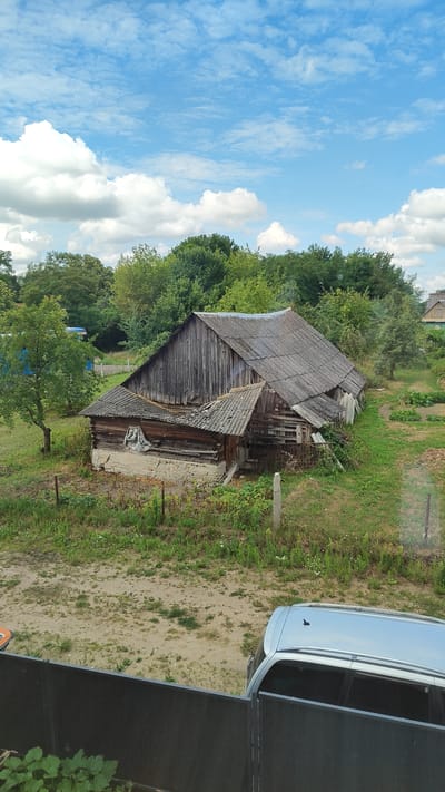 An exterior shot shows a dilapidated wooden barn with a tin roof, surrounded by lush green trees and overgrown grass under a bright blue sky with scattered white clouds. In the foreground, a silver car is parked behind a dark fence. The scene suggests a rural or semi-rural setting, possibly in the Тэльмінскі сельскі Савет region of Belarus, given the architectural style and vegetation. The barn appears to be old and in disrepair, with weathered wooden planks and a partially collapsed roof. The surrounding area is dotted with more trees and some evidence of cultivated land, further reinforcing the impression of a rural environment. The overall mood is one of quiet abandonment and the passage of time.