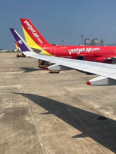 The image depicts an airport tarmac scene in Hòa Cường Ward, Da Nang City, Vietnam, under a clear blue sky. In the foreground, the white wing of an aircraft, possibly an Airbus A320/A321 due to the shape and visible wingtip, extends from the lower left to the middle right, casting a long, distinct shadow onto the concrete surface below.

Positioned in the midground are multiple parked aircraft. The most prominent is a red Vietjet Air Airbus A320/A321, identifiable by its red fuselage with "vietjet Air.com" in white text and "Enjoy Flying!" below. Its tail fin is red with a yellow base and the "Vietjet Air.com" logo. The aircraft registration "VN-A545" is visible near the rear fuselage, and a cargo hatch appears open beneath the "vietjet Air.com" lettering. Yellow ground support equipment, resembling a baggage loader and possibly a pushback tug, is positioned around this red aircraft.

Behind the red Vietjet Air plane, the purple tail fin of another aircraft is visible, displaying a white, stylized 'e' or similar logo. Further back, a portion of a third aircraft's yellow and red tail is faintly discernible. An orange traffic cone stands on the concrete tarmac to the left of the ground support equipment.

In the background, a large, multi-story building complex, likely part of an airport terminal, is visible. Distant hills or mountains are faintly silhouetted against the horizon behind these structures. No individuals are clearly identifiable in the scene.