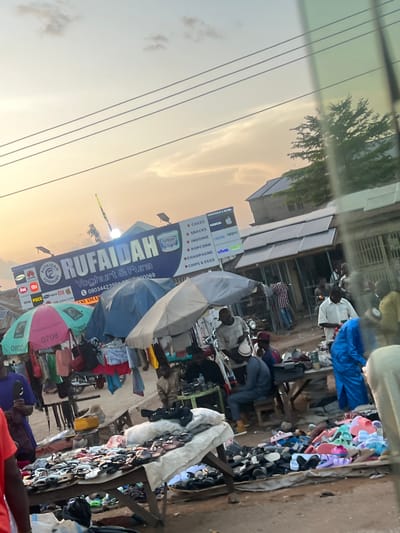 The image depicts an active outdoor market scene during the late afternoon or early evening in Rinjim Gani, Nigeria. The sky displays a gradient of pale orange-yellow at the horizon, transitioning to light blue overhead, with scattered light clouds. Three parallel power lines traverse the upper left sky. A significant greenish-tinted reflection or pane of glass partially obscures and distorts the far right side of the frame.
A prominent blue billboard sign for "RUFAIDAH" is centrally located, advertising "Yoghurt & Fura" and listing "WHOLESALE & RETAIL" products such as "CAKES," "SNACKS," "INDOMIE," "POPCORN," "CHAMPAGNE," and "CHIPS & EGGS." Below the main title, various mobile phone brand logos are displayed, including Samsung, Huawei, itel, Mi, Infinix, POCO, Nokia, Apple, TECNO, oppo, and vivo, alongside a phone number (08034423990). Two solar-powered streetlights are mounted near the billboard.
The market features multiple makeshift stalls, sheltered by large umbrellas; one is distinctively pink and green, while others are blue or off-white/grey. Clothing items, including shirts and shorts, are displayed hanging from racks beneath the umbrellas. In the foreground and mid-ground, a large wooden table covered with a white cloth displays numerous pairs of sandals and slippers in various styles and colors. Adjacent to this, piles of colorful plastic footwear are arranged directly on the unpaved ground.
Numerous individuals are present throughout the market, engaged in commercial activity. Most are men, dressed in casual attire. A man in a white shirt stands on the right side of the image, while another in a blue traditional garment is partially visible further right, obscured by the reflection. On the left-center, a man wearing a grey top and a white cap is seated at a low table. Another individual in a purple shirt is visible on the far left, holding a dark object. The ground is a dry, unpaved surface. In the background, various single-story buildings with corrugated roofs are visible, interspersed with trees.