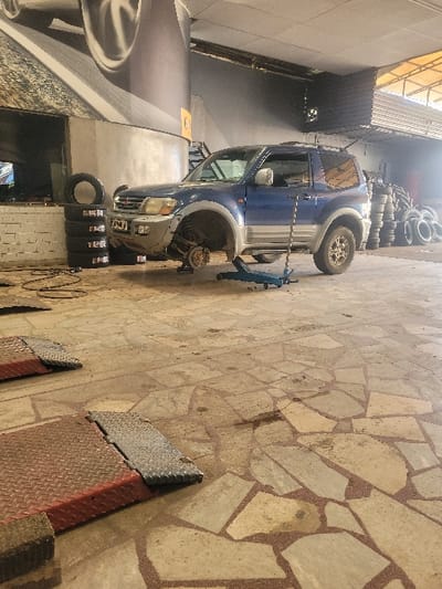 The image depicts an interior scene within a vehicle service or tire shop in Amaliada Municipal Unit, Greece. A two-door, blue and silver Mitsubishi Pajero Pinin/iO SUV is the central subject, elevated with its front-left wheel removed, exposing the wheel hub. The vehicle is supported by a jack stand beneath the front-left frame and a blue hydraulic floor jack positioned under the front-right suspension. To the immediate left of the vehicle, a stack of four tires rests against a low white brick wall, near a large pillar adorned with a graphic of car wheels. In the background on the right, a substantial pile of various tires is visible against a wall or storage area. In the foreground, two red and silver metal vehicle ramps or weighing scales are present on the floor. The floor itself is composed of irregular light-colored stone tiles set in a reddish-brown grout, displaying several dark stains. No individuals are visible within the scene.