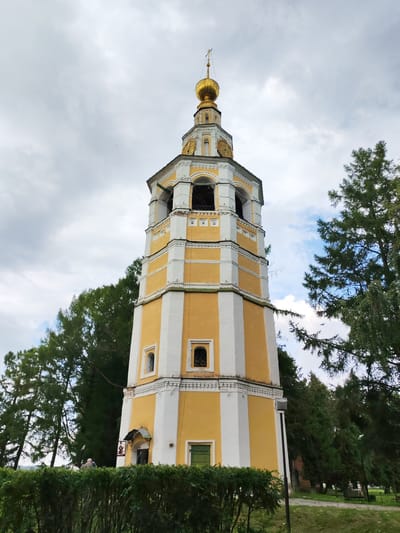 A tall, octagonal bell tower with alternating yellow and white vertical panels stands prominently in the foreground, its golden dome topped with a cross reaching towards a cloudy sky. The tower features arched openings at multiple levels, decorative white trim, and small, barred windows. A green door is visible at the base of the tower, adjacent to a small entrance canopy with an icon. In front of the tower, a dense, dark green hedge lines a path. Beyond the hedge, two individuals are partially visible. Lush green trees surround the bell tower, with some branches extending into the frame from the right. The ground is covered in grass and a paved path. In the background, more trees and what appears to be a fence can be seen, along with a few distant figures and park benches. The overall scene depicts a religious structure within a park-like setting.