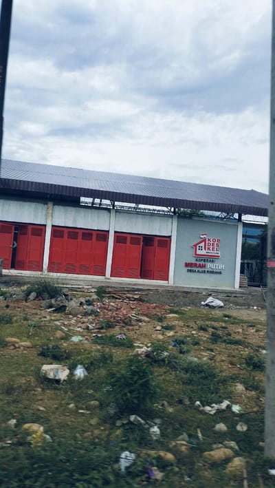 The image displays an exterior view of a building under an overcast sky, likely in Lonbaroh, Indonesia. The building has light grey walls and a dark, possibly corrugated metal roof. The left side of the facade features a series of five prominent red folding doors or shutters. To the right of these doors, a section of the wall serves as a large sign.
The sign prominently features a logo depicting a stylized house with a red and white roof, resembling the Indonesian flag. Above the house, the text "KOP DES KEL" is visible, with "MERAH PUTIH" in smaller red letters underneath. Below the logo, larger red text reads "KOPERASI MERAH PUTIH," and beneath that, in smaller grey letters, "DESA ALUE RINDANG." "Koperasi Merah Putih Desa Alue Rindang" translates to "Red White Cooperative of Alue Rindang Village."
The foreground and midground in front of the building consist of an uneven, unpaved area. This ground is sparsely covered with grass and dirt, and is littered with various debris including rocks, broken bricks, pieces of wood, and scattered plastic waste. This suggests a raw, undeveloped, or possibly neglected area. No individuals or specific activities are visible. The sky is uniformly covered with light grey clouds, indicating an overcast or cloudy day. The overall scene portrays a cooperative building in a somewhat rural or developing setting.